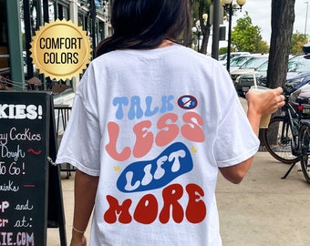 Puede incluir: Camiseta blanca con el texto "Talk Less Lift More" en letras de colores de estilo retro. El texto está en una fuente de burbujas con un esquema de color azul, rosa y rojo.