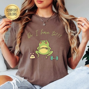 Puede incluir: Camiseta color espresso con un dibujo de una rana de dibujos animados. La rana es verde y amarilla, con los brazos cruzados, y el texto "Do I have to??" está encima. Una pesa y una báscula están al lado de la rana.