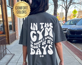 Op de afbeelding: Een donkergrijs T-shirt met de tekst "IN THE GYM IS WHERE I SPEND MOST OF MY DAYS" in wit, met een halterstang symbool onder de tekst.