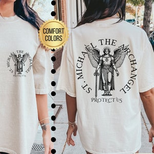 May include: A white t-shirt with a black graphic of an angel with a sword and the text "St. Michael The Archangel Protect Us". The shirt is available in ivory.