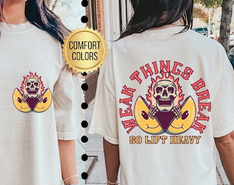 Op de afbeelding: Ivoor kleurig T-shirt met een afbeelding van een schedel in vlammen en een lachend geel ontwerp. De achterkant van het shirt heeft de tekst "WEAK THINGS BREAK SO LIFT HEAVY" in een cirkelvormig ontwerp. Het shirt is een comfort kleur.