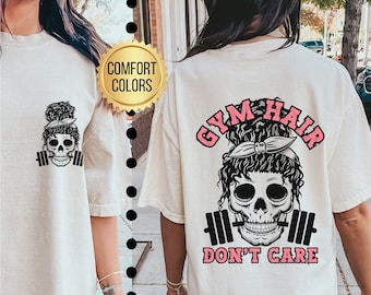 Op de afbeelding: Ivoor kleurig T-shirt met een schedelafbeelding met een hoofdband en halter. De tekst "GYM HAIR DON'T CARE" staat in het roze op de achterkant. Het shirt is gemarkeerd met "Comfort Colors".