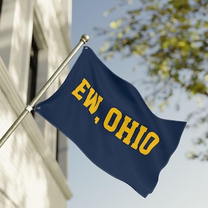 Könnte beinhalten: Eine marineblaue Flagge mit dem gelben Schriftzug "EW, OHIO". Die Flagge ist an einer silbernen Stange befestigt und weht im Wind, vor dem Hintergrund eines Gebäudes und eines blauen Himmels.