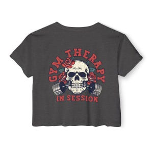 May include: Dark gray cropped t-shirt with a skull graphic wearing a barbell in its mouth. The skull is surrounded by red roses and the text "Gym Therapy In Session".