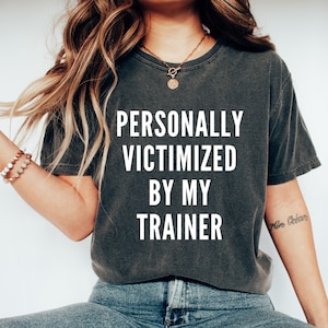 Pode incluir: T-shirt cinza escuro com o texto "PERSONALLY VICTIMIZED BY MY TRAINER" em letras brancas. A camisa tem uma aparência ligeiramente desbotada e um ajuste folgado. A modelo está usando jeans azuis.