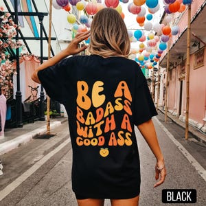Könnte beinhalten: Schwarzes T-Shirt mit dem Text "BE A BADASS WITH A GOOD ASS" in orange und gelben Buchstaben. Unter dem Text befindet sich eine kleine orangefarbene Pfirsichgrafik. Das Shirt ist ein lässiger Stil.