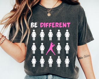May include: A dark gray t-shirt with the text "BE DIFFERENT" printed in white. The shirt features a graphic of a pink female figure playing baseball surrounded by white female figures.