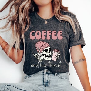 May include: A dark gray t-shirt with the text "COFFEE and hip thrust" in pink lettering. A cartoon skeleton wearing a pink beanie is holding a pink coffee cup.