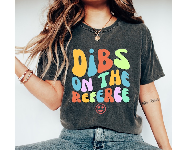 May include: A dark gray t-shirt with the text "Dibs on the Referee" in colorful, retro-style lettering. A smiley face is below the text.