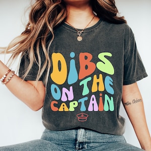 May include: A dark grey t-shirt with the text "DIBS ON THE CAPTAIN" in colourful, retro-style lettering. A small captain's hat icon is below the text.