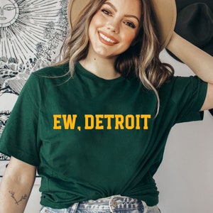 May include: A dark green t-shirt with the text "EW, DETROIT" in yellow block letters. The shirt is a crew neck style and appears to be made of a soft material. The overall theme is casual wear.