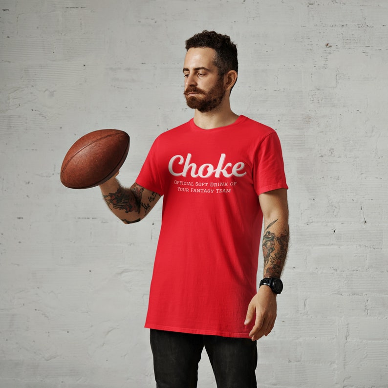 Pu&ograve; includere: Un uomo indossa una maglietta rossa con il testo "Choke Official Soft Drink of Your Fantasy Team" in bianco. Sta tenendo un pallone da football marrone.