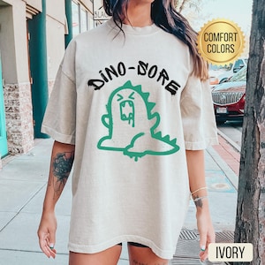 Puede incluir: Una camiseta de gran tamaño de color beige claro con un gráfico de dinosaurio de dibujos animados verde y el texto "Dino-Sore" en negro. La camiseta está hecha de una tela suave y cómoda y tiene un corte holgado.