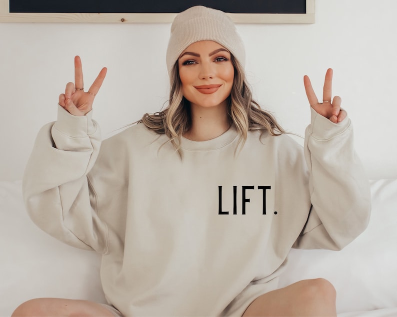 May include: A beige sweatshirt with the word "LIFT." printed in black on the front. The model is wearing a beige beanie and is making a peace sign with both hands.