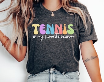 May include: A dark grey t-shirt with the text "TENNIS is my favourite season" in colourful, rounded letters. The letters are in shades of yellow, purple, blue, pink, and green.