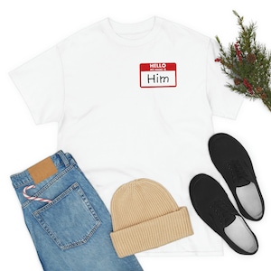 Pode incluir: Camiseta branca com uma etiqueta de nome vermelha que diz "HELLO MY NAME IS Him". A camiseta está deitada sobre um fundo branco com um par de sapatos pretos, um gorro e calças de ganga azuis.