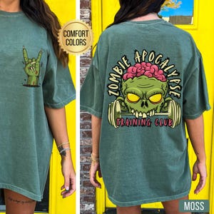 May include: A moss green t-shirt with a zombie-themed design. The front shows a green zombie hand making a rock and roll gesture. The back has a zombie head lifting weights with the text "ZOMBIE APOCALYPSE TRAINING CLUB".