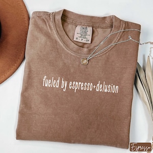 May include: A brown Comfort Colors t-shirt with the phrase "fueled by espresso+delusion" in white script. A silver necklace with a small pendant rests on the shirt. A brown hat is visible in the upper left corner.