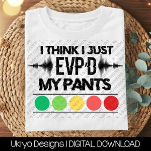 May include: A white t-shirt featuring the black text I THINK I JUST EVP'D MY PANTS with a central sound wave graphic. Below the text are five horizontal circles in green, light green, yellow, orange, and red, framed by two black lines.