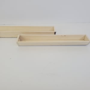 May include: Two unfinished, rectangular wooden trays. The trays are light-colored with a natural wood grain. One tray is slightly smaller than the other. The edges of the trays are angled, and the bottoms are flat. The trays are empty.