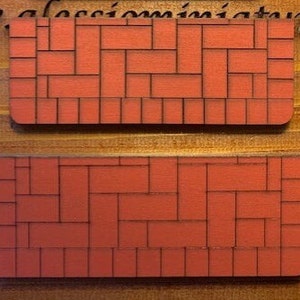 May include: Two rectangular, red wooden miniature brick wall panels with black grout lines. The panels are arranged in a staggered brick pattern. The background is a wooden surface.