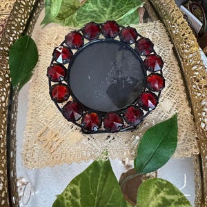 May include: A black metal candle holder with a circular design, adorned with vibrant red gemstone accents. The candle holder sits on a decorative lace doily within a gold-framed tray, complemented by green leaves.