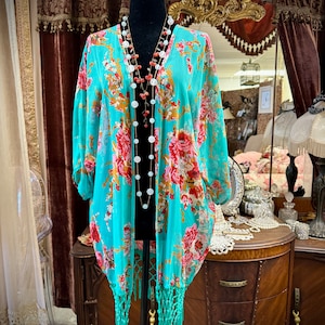 May include: A turquoise floral print kimono with a long fringe hem. The kimono is on a mannequin and has a long beaded necklace.