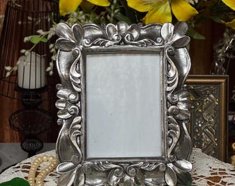 May include: Ornate silver-toned picture frame with floral and scroll detailing. The frame has a rectangular opening and sits on a lace doily. A strand of faux pearls and green leaves are draped nearby. Yellow flowers are in the background.