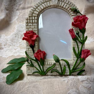 May include: An arched picture frame with a brick-like design, adorned with red rose blossoms and green leaves. The frame has a neutral color palette, with a white interior for the photo.