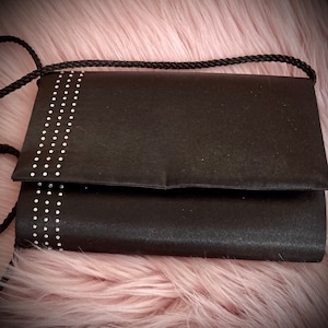May include: A black satin clutch bag with a silver rhinestone design. The bag has a long strap.