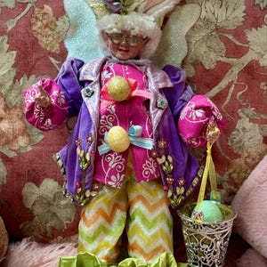 May include: A whimsical Easter bunny figurine with large ears, wearing a purple coat, pink shirt, and patterned pants. The bunny holds a small silver basket with an egg. The outfit is adorned with floral and jewel accents. The figurine has glasses and a smiling face.