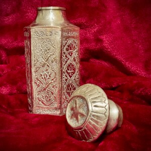 May include: A silver octagonal bottle with an ornate design and a matching lid. The bottle is sitting on a red velvet surface.
