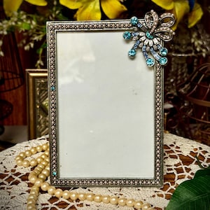 May include: A silver picture frame with a blue and white jeweled embellishment on the top right corner. The frame is sitting on a white crocheted doily.