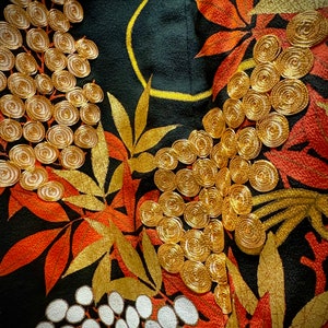 May include: Close-up of a black fabric with gold and orange embroidery. The design features stylized leaves and flowers with intricate details. The fabric has a textured surface.