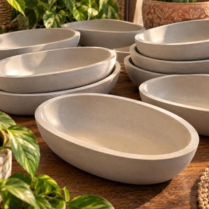 May include: A collection of multiple light gray oval-shaped bowls with smooth, matte surfaces. The bowls are arranged in a stacked and overlapping formation, showcasing their deep, rounded interiors and thick, uniform rims in a neutral tone.