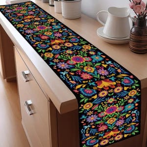 May include: A black table runner with a vibrant floral pattern. The design features colorful flowers in shades of pink, blue, yellow, and orange against a black background. The runner is rectangular and placed on a wooden surface.