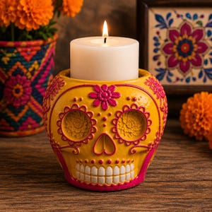 May include: A yellow ceramic skull candle holder featuring bright pink floral patterns around the eye sockets and forehead, with a white pillar candle burning inside. The skull has a detailed row of white teeth and pink accents along the base.