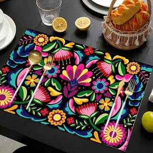 May include: A black placemat with a vibrant floral design in pink, yellow, blue, and green. Gold-colored fork and spoon are on the placemat. A white cup and plate, a glass, and a basket of bread are also on the table.