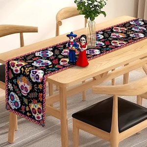 May include: A wooden dining table with a black and colorful Day of the Dead skull table runner. Two figurines and a vase with greenery are on the table. The table is surrounded by wooden chairs with black seats.