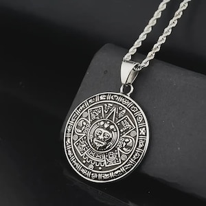 May include: Silver-colored pendant necklace featuring a detailed Aztec calendar design. The circular pendant has intricate carvings and a central face. The necklace chain is a twisted rope style, and the pendant hangs against a dark background.