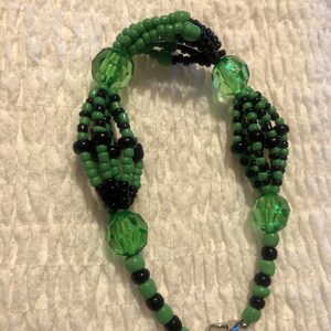 K&ouml;nnte beinhalten: Ein gr&uuml;nes und schwarzes Perlenarmband mit einem silbernen Verschluss. Das Armband ist aus kleinen Perlen und gr&ouml;&szlig;eren gr&uuml;nen Glasperlen gefertigt.