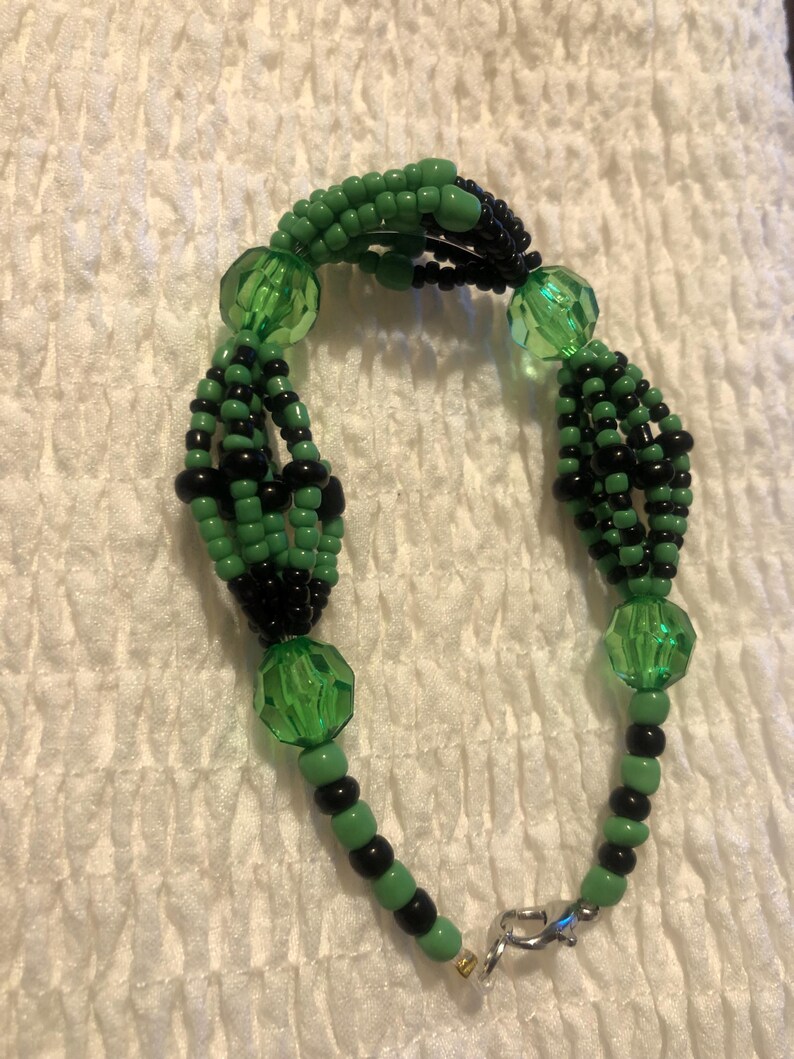 K&ouml;nnte beinhalten: Ein gr&uuml;nes und schwarzes Perlenarmband mit einem silbernen Verschluss. Das Armband ist aus kleinen Perlen und gr&ouml;&szlig;eren gr&uuml;nen Glasperlen gefertigt.