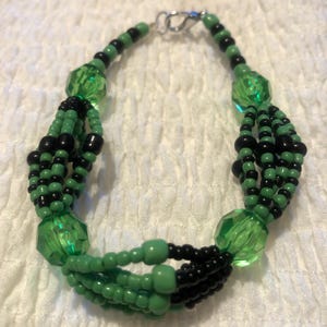 May include: A green and black beaded bracelet with a silver clasp. The bracelet is made with multiple strands of beads, including small round beads and larger faceted beads.