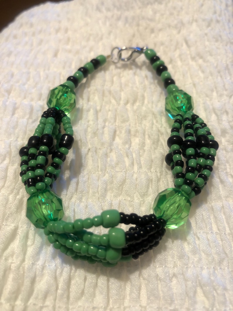 K&ouml;nnte beinhalten: Ein gr&uuml;nes und schwarzes Perlenarmband mit einem silbernen Verschluss. Das Armband besteht aus mehreren Perlenreihen, darunter kleine runde Perlen und gr&ouml;&szlig;ere facettierte Perlen.
