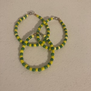 May include: Three beaded bracelets. Each bracelet is made of alternating yellow and green beads. The bracelets are connected with silver-colored clasps. The bracelets are on a light-colored surface.