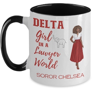 May include: White ceramic coffee mug with a black handle and black rim. The mug has a red graphic that says "DELTA Girl in a Lawyer World" with a small elephant illustration. The graphic also includes the text "SOROR CHELSEA".