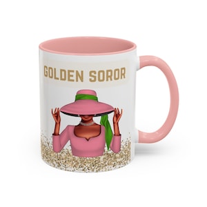 Puede incluir: Taza de cerámica blanca con interior y asa de color rosa. La taza presenta una inscripción dorada que dice "GOLDEN SOROR" y un gráfico de una mujer con un sombrero rosa con una cinta verde y un vestido rosa. El fondo es un patrón de purpurina dorada.