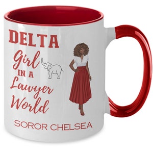 May include: White ceramic coffee mug with a red rim and handle. The mug features a red graphic of a woman in a red skirt and a white top, with the text "DELTA Girl in a Lawyer World SOROR CHELSEA".