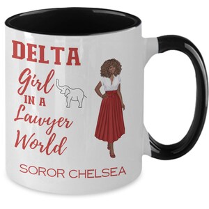 May include: White ceramic coffee mug with a black rim and handle. The mug has a red graphic that says "DELTA Girl in a Lawyer World" with a small elephant illustration. The graphic also includes the text "SOROR CHELSEA".