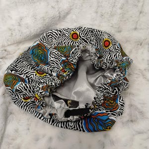 May include: A patterned hair bonnet with a zebra stripe design in black and white, accented with orange, blue, and yellow circular patterns. The interior is a silver satin material. The bonnet is designed to protect hair.